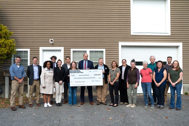 A large group of people hold an oversized check in front of a brownish semi-industrial building.